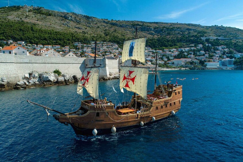 A wooden ship with sails on a calm sea, a coastal town in the background, and a hilly landscape. 1