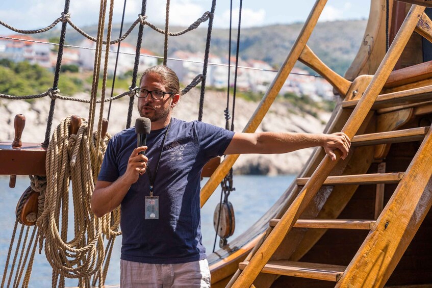 A person standing on a wooden ship deck, holding a microphone, with ropes and a wooden ladder in the background. 4