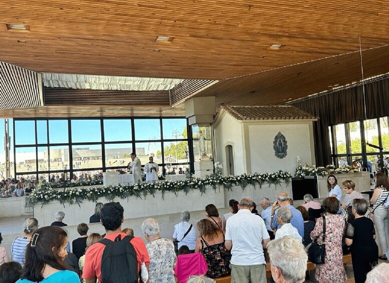 A large gathering of people in an open-air venue with a stage, wooden ceiling, and a view of buildings in the background. 5
