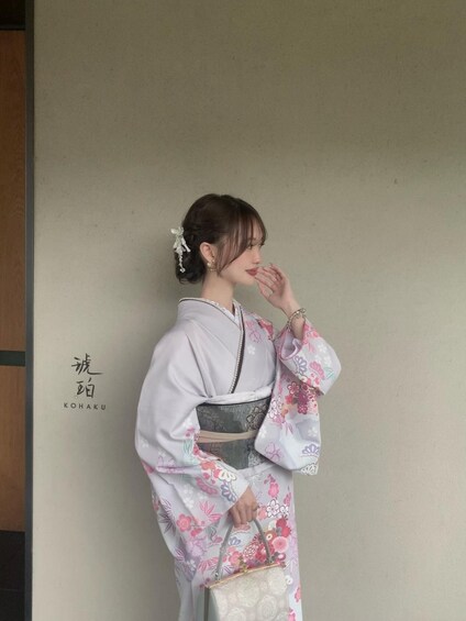 A person in traditional attire with a floral kimono, holding a handbag, standing against a plain wall with a sign. 2