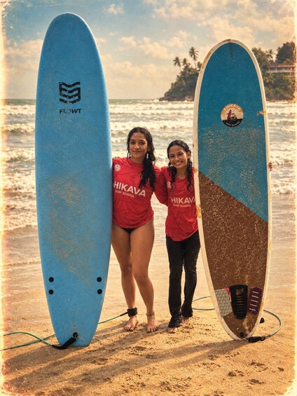 Two people holding surfboards on a beach with palm trees and a clear sky in the background. 2