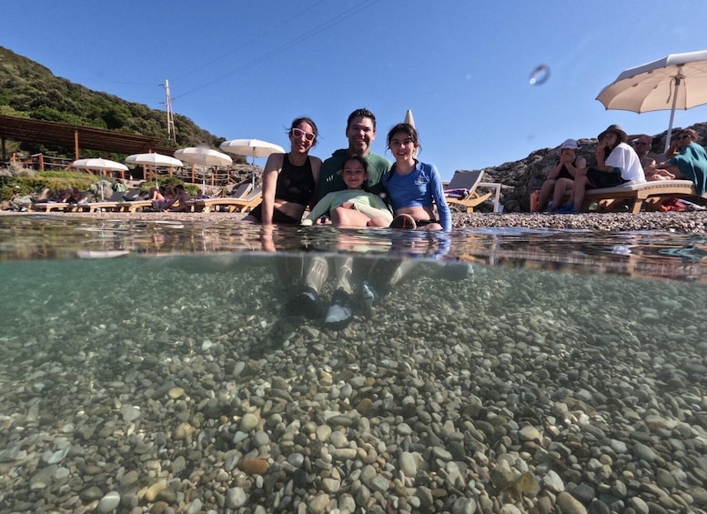 A family in a clear shallow pool with pebbles, surrounded by lounge chairs and umbrellas on a sunny beach. 3