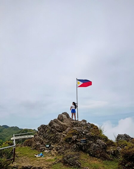 A person standing on a rocky peak with a flag, surrounded by greenery and a cloudy sky. 2