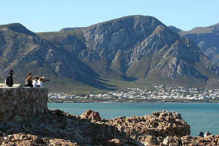 A rocky coastline with people sitting on a ledge overlooking a town and mountains in the background. 2