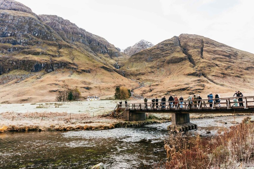 A bridge over a river with people walking on it, surrounded by mountains and a grassy landscape. 4