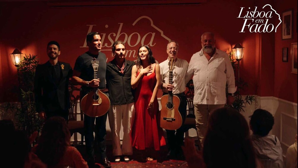 A group of people on a stage with musical instruments, a red wall with text, and a logo in the corner. 2