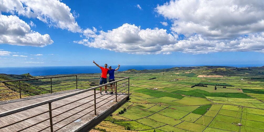 Two people standing on a wooden platform with a scenic view of green fields and the ocean. 1