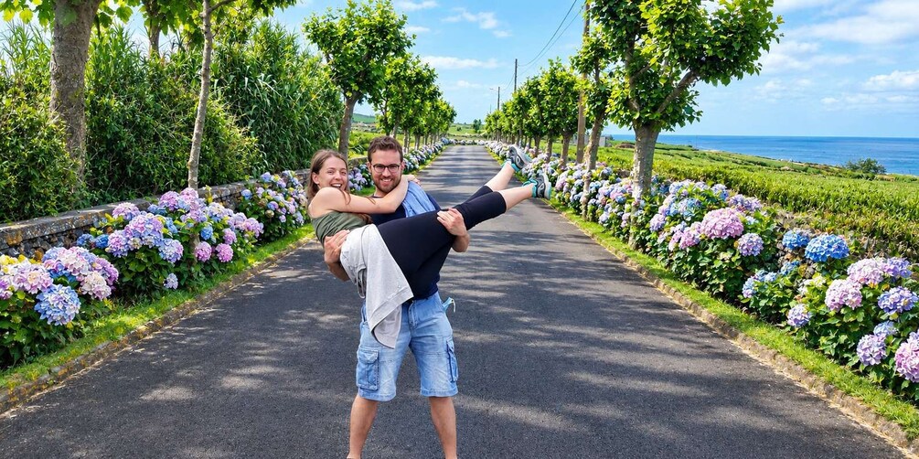A couple embracing on a tree-lined road with hydrangea flowers and a view of the ocean. 3