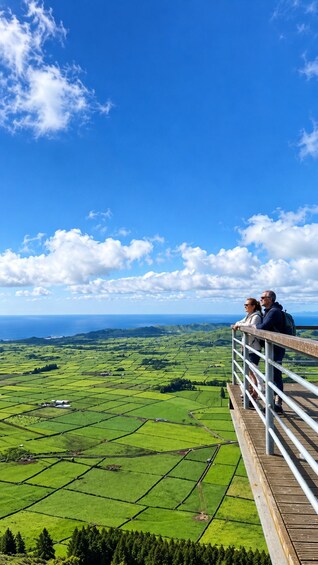 A scenic view from a wooden platform overlooking a green landscape with fields and a distant body of water under a clear blue sky. 5