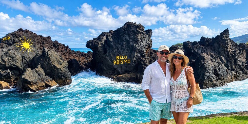 A couple standing by a rocky coastline with waves crashing against the rocks. The text 'BELLO ARISMIO' is visible on one of the rocks. 4