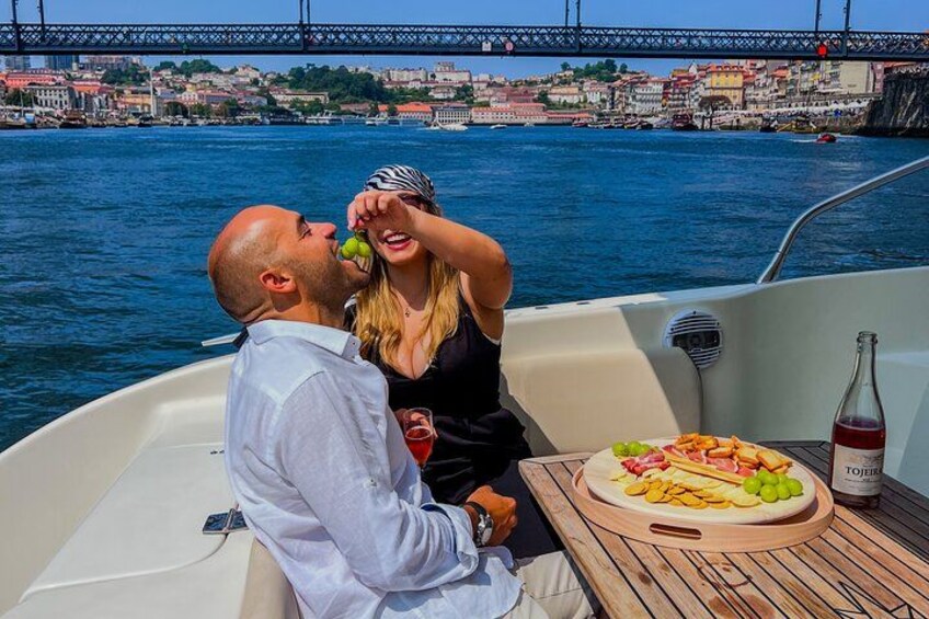 A couple on a boat with a table of food and drinks, a bottle of wine, and a cityscape with a bridge in the background. 4