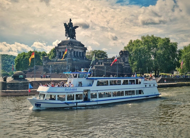 A white and blue boat named 'Goldstück' on a river, with a statue and flags in the background. 1