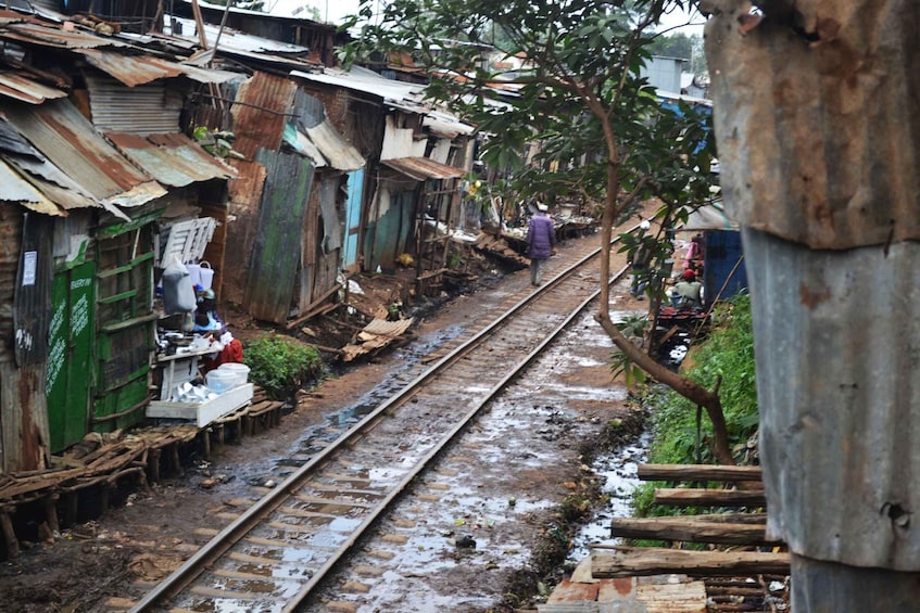 A railway track with a person walking away, surrounded by makeshift structures and greenery. 5
