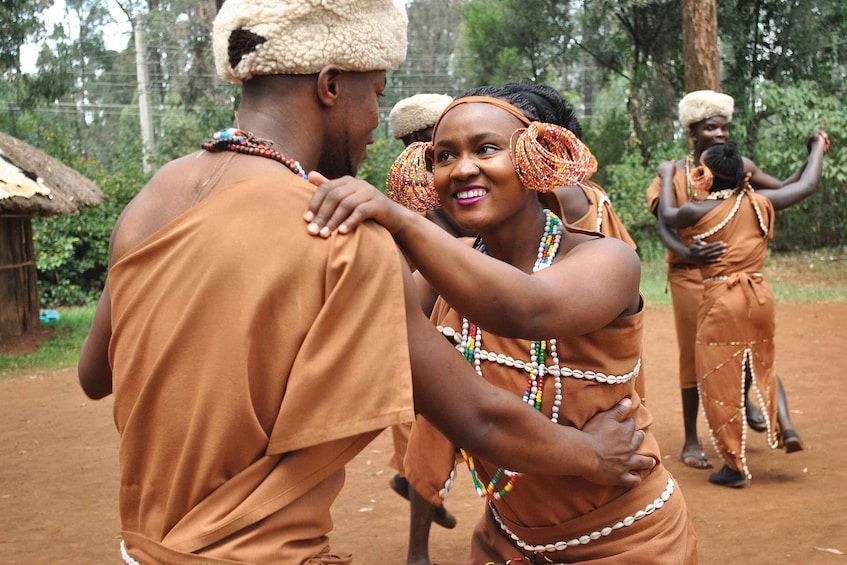 People in traditional attire, one with a head covering, in an outdoor setting with trees and a thatched-roof structure. 4
