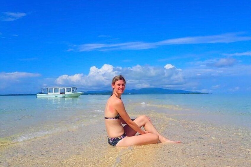 A person sitting on a beach with clear water and a boat in the background. 4