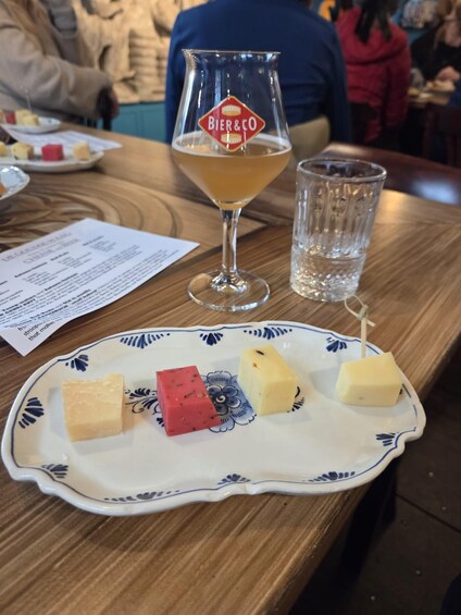 A glass of beer with a logo, a plate with cheese cubes, and a menu on a wooden table. 2