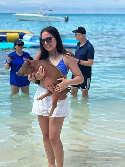 A woman holding a piglet on a beach with clear water and boats in the background. 4
