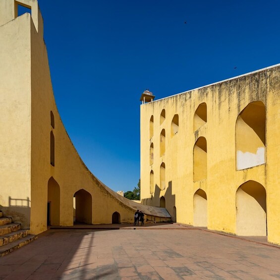 A courtyard with yellow walls and arches, a clear blue sky, and a few people walking. 1