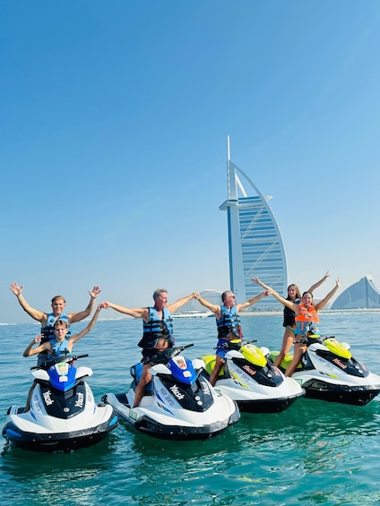 Four people on jet skis with arms raised, water sports gear, and a sail-shaped building in the background. 3