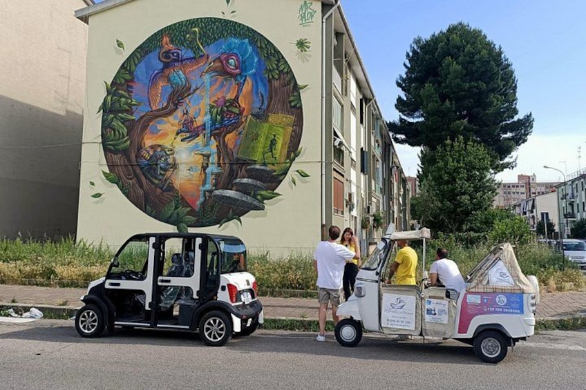 A mural on a building, a small vehicle with people, and a large tree. 2