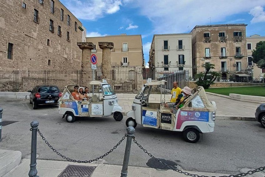 A white tour vehicle with passengers, parked on a street with buildings and a no parking sign in the background. 3