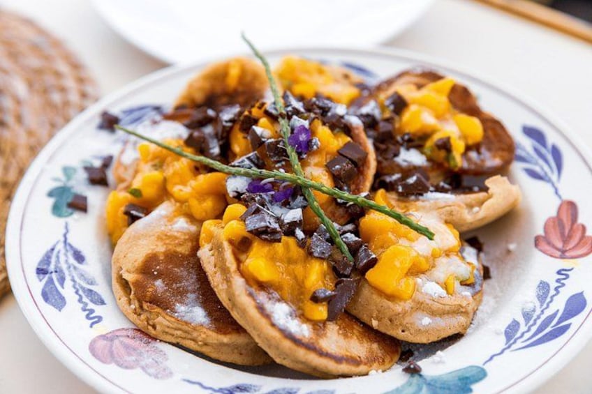 A plate of pancakes topped with yellow sauce, chocolate pieces, and a sprig of herb. 3