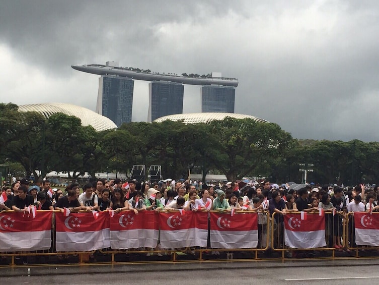 Suuri ihmisjoukko heiluttaa Singaporen lippuja suuren rakennuksen edessä. 3