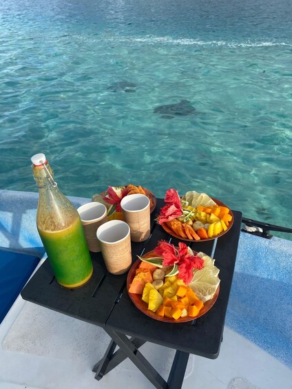 A table with a bottle, cups, and a fruit platter by a clear blue sea. 1
