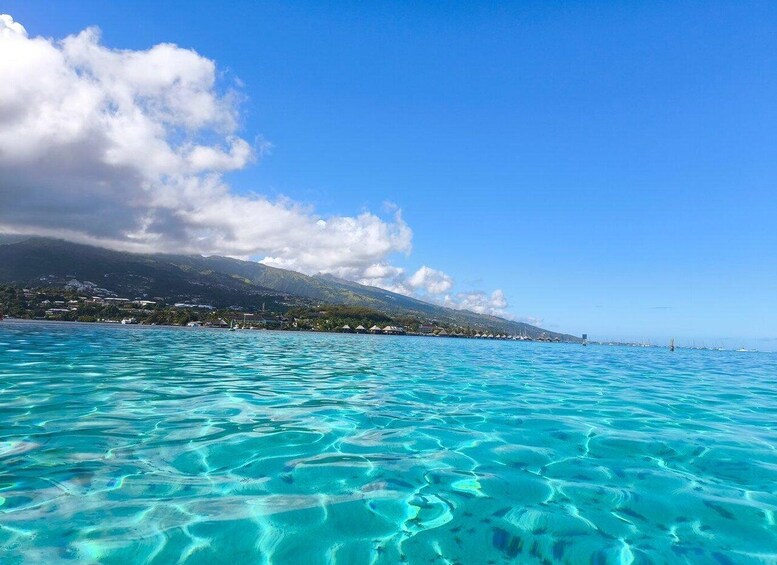Clear turquoise waters, a distant shoreline with buildings, and a mountain range under a partly cloudy sky. 3