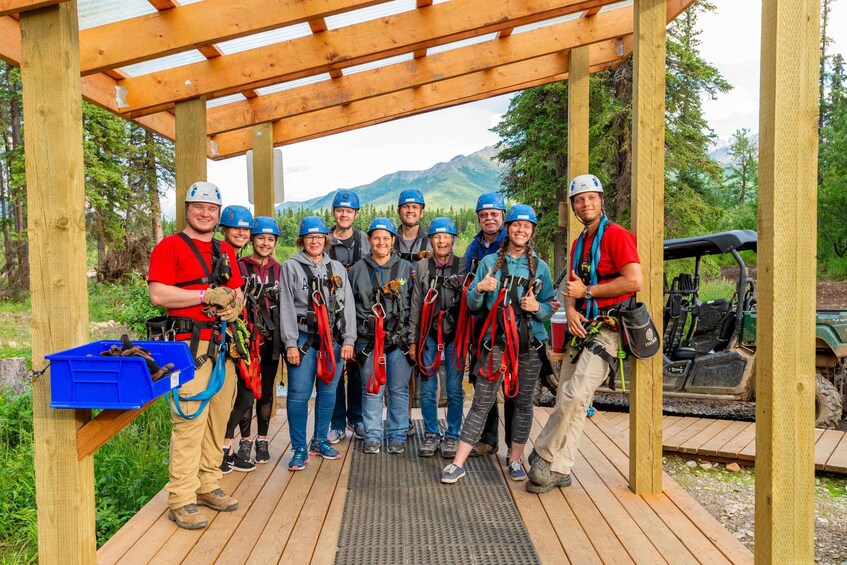 A group of people in helmets and harnesses standing under a wooden structure with a mountain backdrop. 5