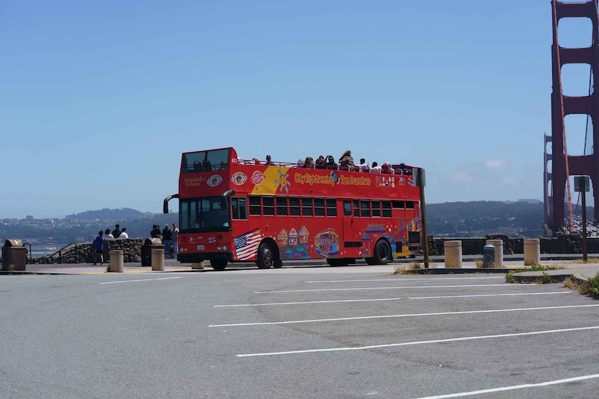 A red double-decker bus with 'CitySightseeing San Francisco' written on it, parked in a parking lot with the Golden Gate Bridge in the background. 2
