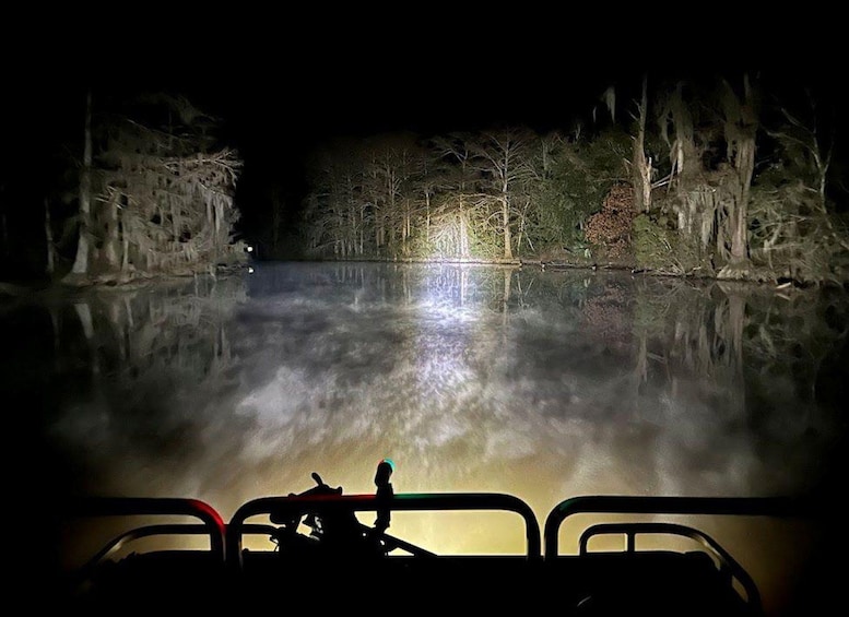 A nighttime scene with a calm river reflecting lights, surrounded by trees and a distant illuminated structure. 1