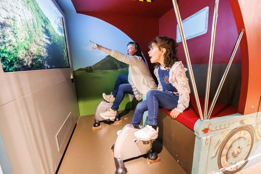 Two people on stationary bikes facing a large screen displaying a landscape. 4