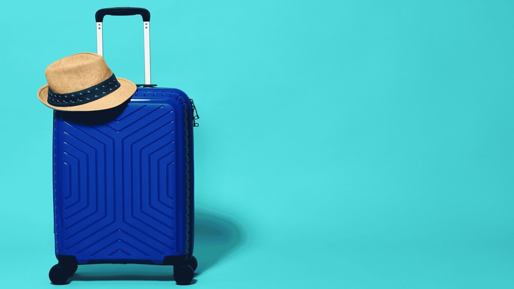A blue suitcase with a straw hat on top, standing against a teal background. 5