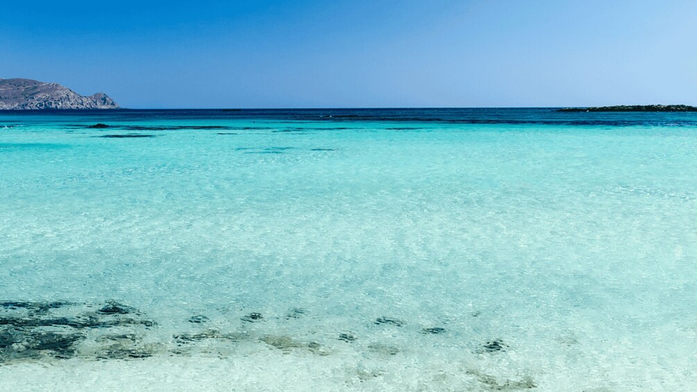 A clear turquoise sea with visible underwater rocks and a distant rocky island. 3