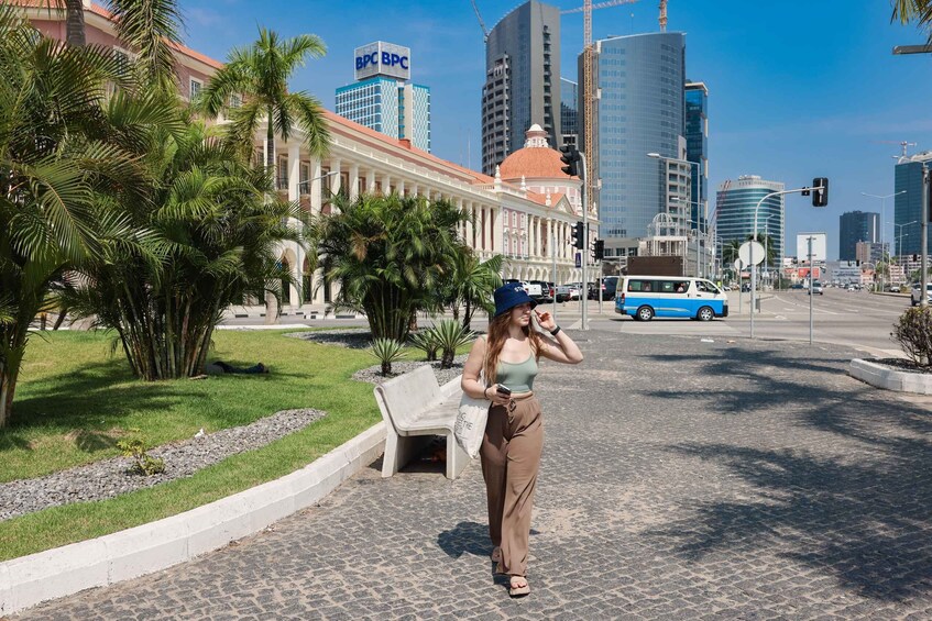 A woman walking on a city street with palm trees, a white bench, and modern buildings in the background. 2