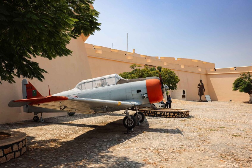 A vintage aircraft parked in a courtyard with a stone floor, surrounded by trees and a building with a flat roof. 3