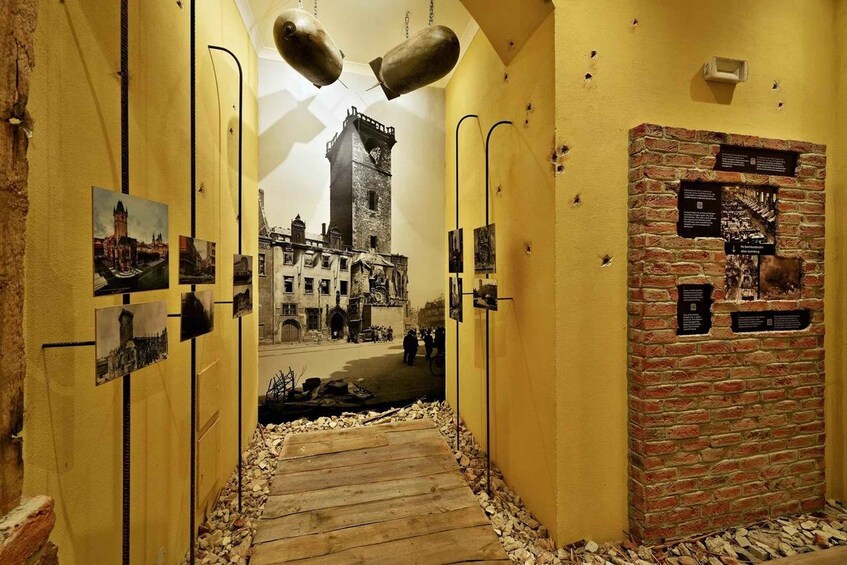 A corridor with yellow walls, wooden floor, and pictures of buildings. There are two large metal objects hanging from the ceiling. 5