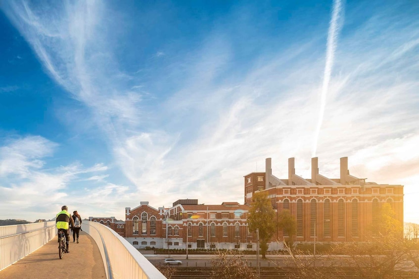A bridge with cyclists, a brick building with chimneys, and a clear sky. 2