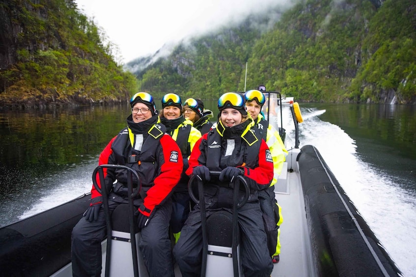 A group of people in red and black jackets on a boat, surrounded by a river and forested hills. 1