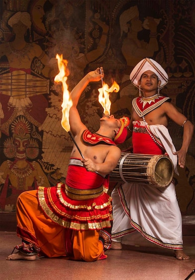 A person performing a fire act with flames, another person playing a drum, and a detailed tapestry in the background. 1