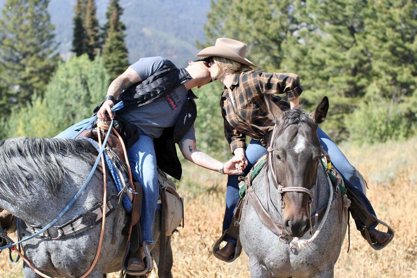 Two people on horseback in a natural setting with trees and a mountainous background. 4