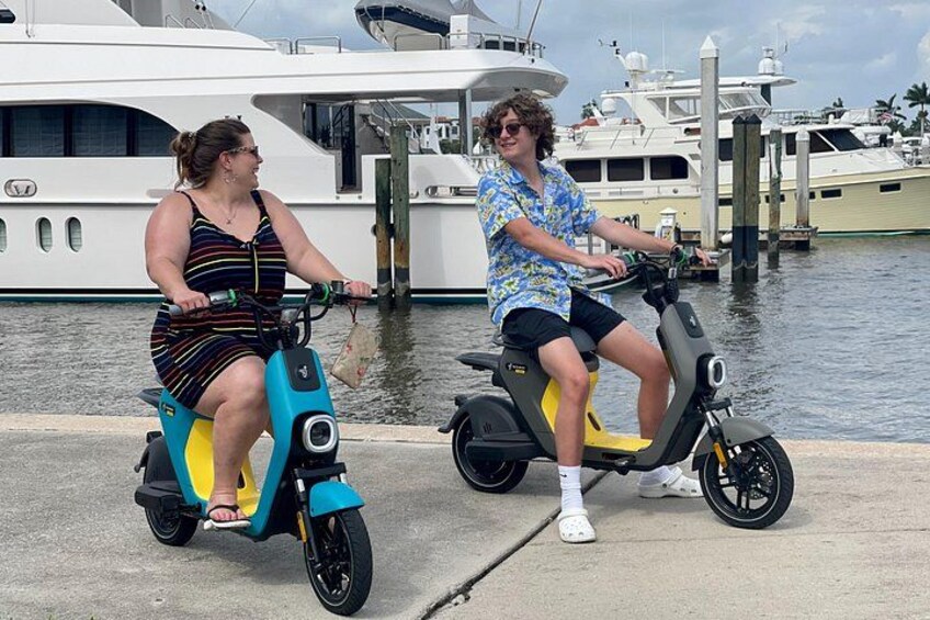 Zwei Personen fahren auf Rollern in der Nähe eines Yachthafens mit Booten und Stegen. 3