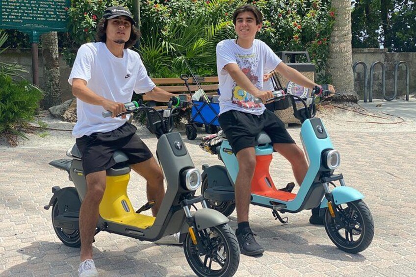 Zwei Personen fahren auf E-Scootern auf einem asphaltierten Weg, im Hintergrund grüne Vegetation. 2