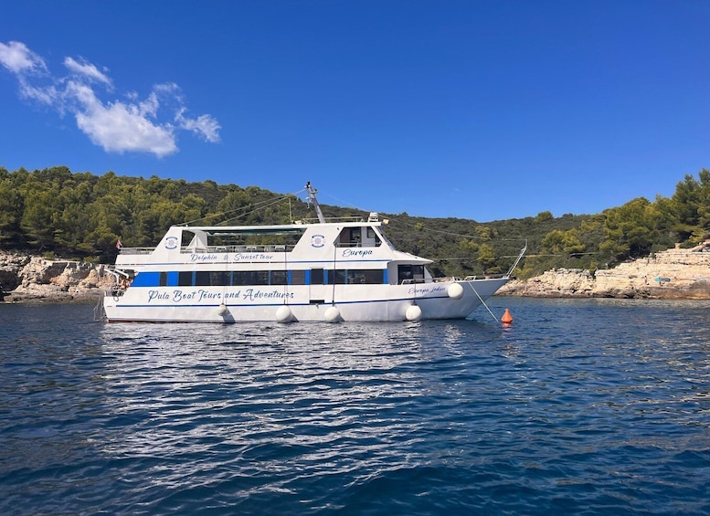 A white boat named 'Europa' with 'Pula Boat Tours and Adventures' written on its side, docked near a rocky shoreline with trees. 1