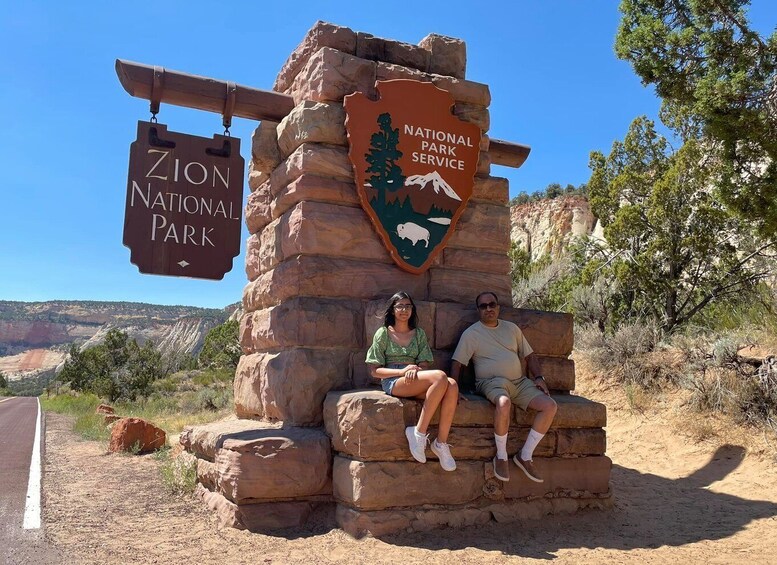 A Zion National Park sign with a woman and a man sitting on a rock structure. 2
