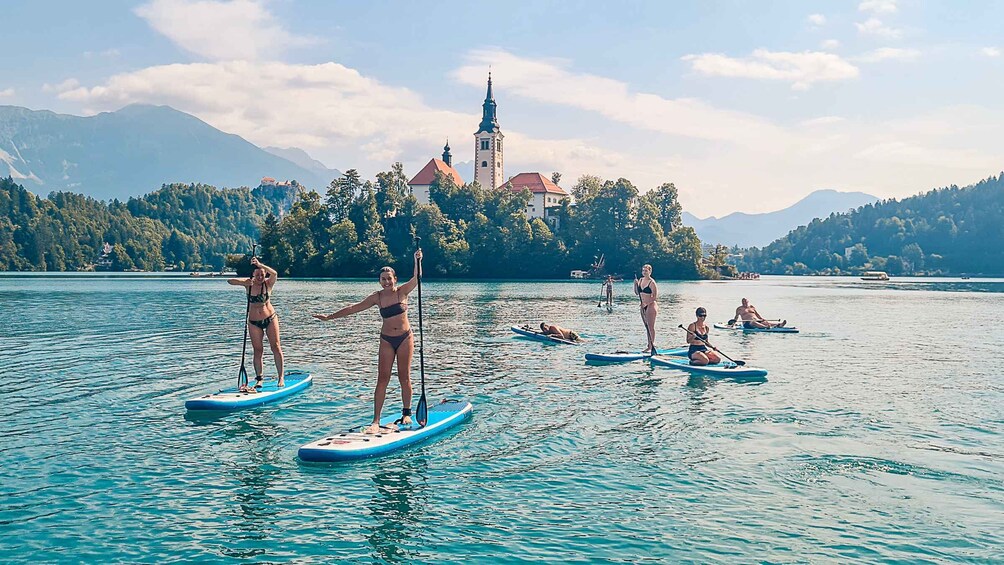People paddleboarding on a calm lake with a church and mountains in the background. 1