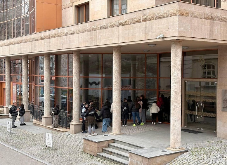 Een gebouw met grote glasramen en kolommen, mensen staan buiten. 3
