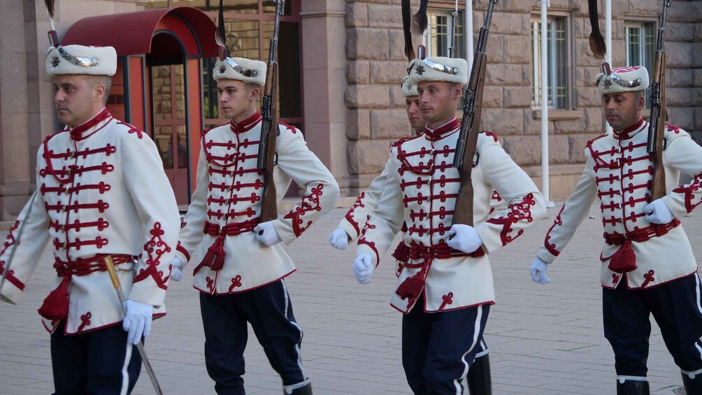 Un grupo de guardias uniformados con atuendo ceremonial y rifles, formados en un área pavimentada. 5