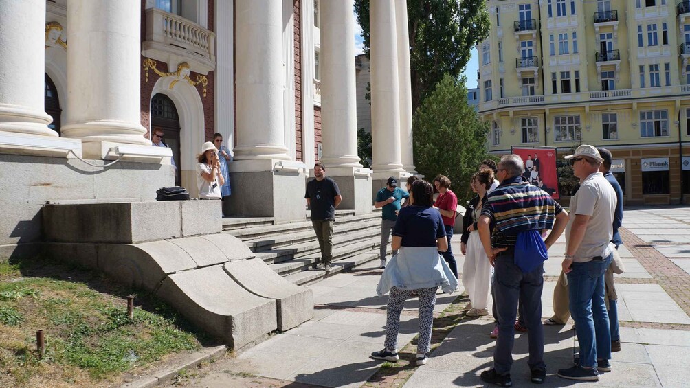 A group of people standing in front of a building with columns and a statue. 2